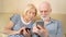 Cheerful good-looking senior couple sitting on sofa at home. Using smartphone, browsing, reading