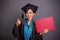 Cheerful graduated girl thumb up while holding placard, portrait on grey background