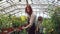 Cheerful greenhouse worker is watering plants in workplace with her helpful daughter and talking to child while little