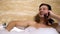 Cheerful guy laughing, talking on phone while taking bath with foam, relaxation