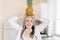 Cheerful happy pretty retired Caucasian woman in white shirt, holding a pineapple on her head, looking at camera and