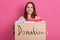 Cheerful helpful volunteer standing isolated over pink background in studio, holding box with inscription Donation, full of