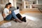 Cheerful international millennial couple looking at laptop, have video call, enjoy coffee cup, sit on floor