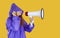 Cheerful joyful woman in waterproof purple raincoat with hood makes announcement using megaphone.