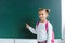 cheerful kid smiling while pointing with finger at green chalkboard .