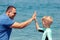 Cheerful little boy in surfing swimwear giving high five to dad on beach expressing trust and confidence