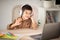 Cheerful little boy in wireless headphones sits at table, studies, has video call on laptop, waving hand in room