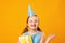 Cheerful little girl celebrates birthday. A child in a blue dress and cap holds a box with a gift. Closeup portrait on yellow