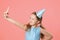 Cheerful little girl celebrates birthday. The child holds the phone, takes a selfie. Closeup portrait on pink coral background