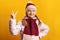 Cheerful little girl in a jacket, scarf and hat on a yellow background. The child shows a victory sign. Success concept