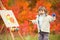 Cheerful little girl in a jump with a brush and paint, draws in the park of autumn leaves, painting of the Little Child