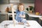 A cheerful little girl, soiled in flour, is sitting on the table, laughing and holding a rolling pin