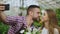 Cheerful loving couple gardeners taking selfie picture on smartphone camera and kissing while working in greenhouse