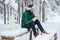 Cheerful male enjoys communication on smart phone and loneliness, calm atmosphere, sits on wooden bench covered with snow in winte