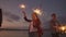 Cheerful male and female friends are running along the beach at sunset, holding sparkling fireworks and runaway lights