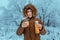 Cheerful man in a down jacket with a hood drinks hot tea from a thermos against the background of frosted trees after a cold