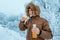 Cheerful man in a down jacket with a hood drinks hot tea from a thermos against the background of frosted trees after a cold