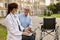 Cheerful mature recovering patient with wheelchair having conversation with young female doctor working on a laptop in