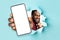 Cheerful millennial african american guy with beard look through hole in blue paper and pointing finger at smartphone