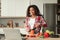 Cheerful millennial african american lady cuts organic vegetables and looks at laptop, enjoys cooking food