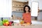 Cheerful millennial black lady cuts organic vegetables at salad, shows smartphone with blank screen in kitchen