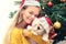 Cheerful millennial girl with santa hat embracing her gift little bichon frise dog under Christmas tree