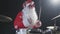 Cheerful musician dressed as Santa Claus plays drums on a black studio backlit background. A man in a red festive suit
