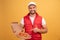 Cheerful pizza delivery man stands with carton boxes, point to the pizza, wears yellow hat and white t shirt, smiling during