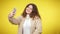 Cheerful plus-size millennial woman taking selfie on smartphone at yellow background. Device addicted young charming