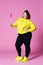 Cheerful plus size model in casual clothes with big lollipop, fat woman in black jeans and yellow jumper on pink background