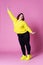 Cheerful plus size model in casual clothes, fat woman in black jeans and yellow jumper on pink background