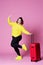 Cheerful plus size model with red suitcase, fat woman in black jeans and yellow jumper on pink background