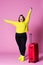 Cheerful plus size model with red suitcase, fat woman in black jeans and yellow jumper on pink background