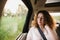 Cheerful positive curly young woman in casual wear sitting in automobile backseat with fastened seatbelt and looking away