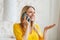 Cheerful pretty young blond european female in yellow clothes speaks by phone, gesticulates, hold hand with empty space