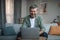 Cheerful retired european male with beard blogger typing on computer or watching video in living room