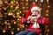 Cheerful santa helper girl with cup of tea sitting indoor near decorated xmas tree with lights, dressed in red sweater - Merry