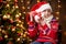Cheerful santa helper girl with gift box sitting indoor near decorated xmas tree with lights, dressed in red sweater - Merry