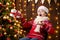 Cheerful santa helper girl with gift box sitting indoor near decorated xmas tree with lights, dressed in red sweater - Merry