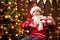 Cheerful santa helper girl with gift box sitting indoor near decorated xmas tree with lights, dressed in red sweater - Merry