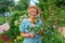 Cheerful senior lady in glasses and a hat stands in the garden among the flowers and holds a raspberry branch. Cover portrait for