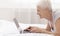 Cheerful senior lady typing on laptop computer, lying in bed