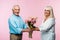 cheerful senior man giving bouquet with flowers to happy retired wife on pink.