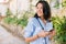 Cheerful shot portrait of happy attractive woman with mobile phone looking at one side and waiting friends on the city street.