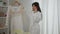 Cheerful smiling Middle Eastern bride talking on the phone holding hanger with wedding dress. Positive happy young woman