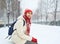 Cheerful smiling woman in white down jacket and red cap, scarf and mittens sitting on a bench in the alley on the snowy street