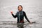 cheerful sporty woman in wetsuit with vest standing in the water and shows hand gesture.