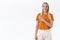 Cheerful stylish blond caucasian woman in orange t-shirt smiling enthusiastic, pointing finger left at blank white space