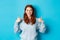 Cheerful teen girl with red hair, showing thumbs up in approval, like and praise gesture, standing over blue background