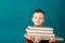 Cheerful thoughtful little school boy in school uniform with backpack and big pile of books standing against blue wall. Looking